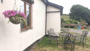 Un bâtiment blanc avec un pot de fleurs suspendu près d’une fenêtre, une terrasse avec des chaises et une table en métal, et un terrain de jeux en arrière-plan.