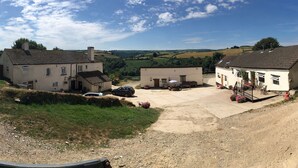 Un cadre rural avec deux bâtiments blancs, dont l’un possède un porche et des sièges à l’extérieur, et un chemin de terre menant à une voiture.