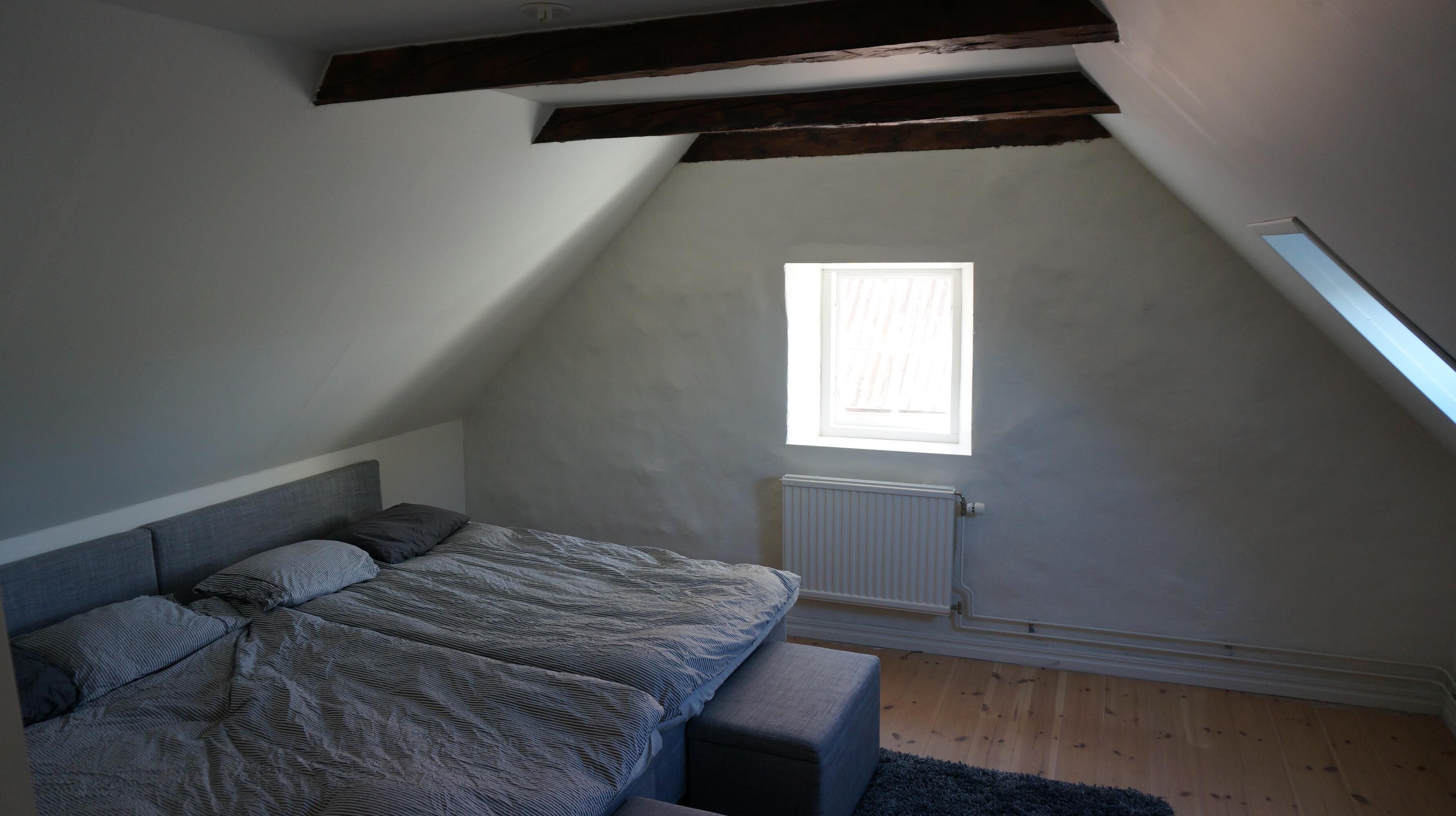 Una camera da letto con soffitto spiovente, una finestra, un termosifone, un letto con coperta a righe e una panca.