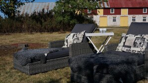 Dos sillones de mimbre con cojines negros y almohadas estampadas, ubicados en un césped con una cerca de metal y un edificio rojo al fondo.