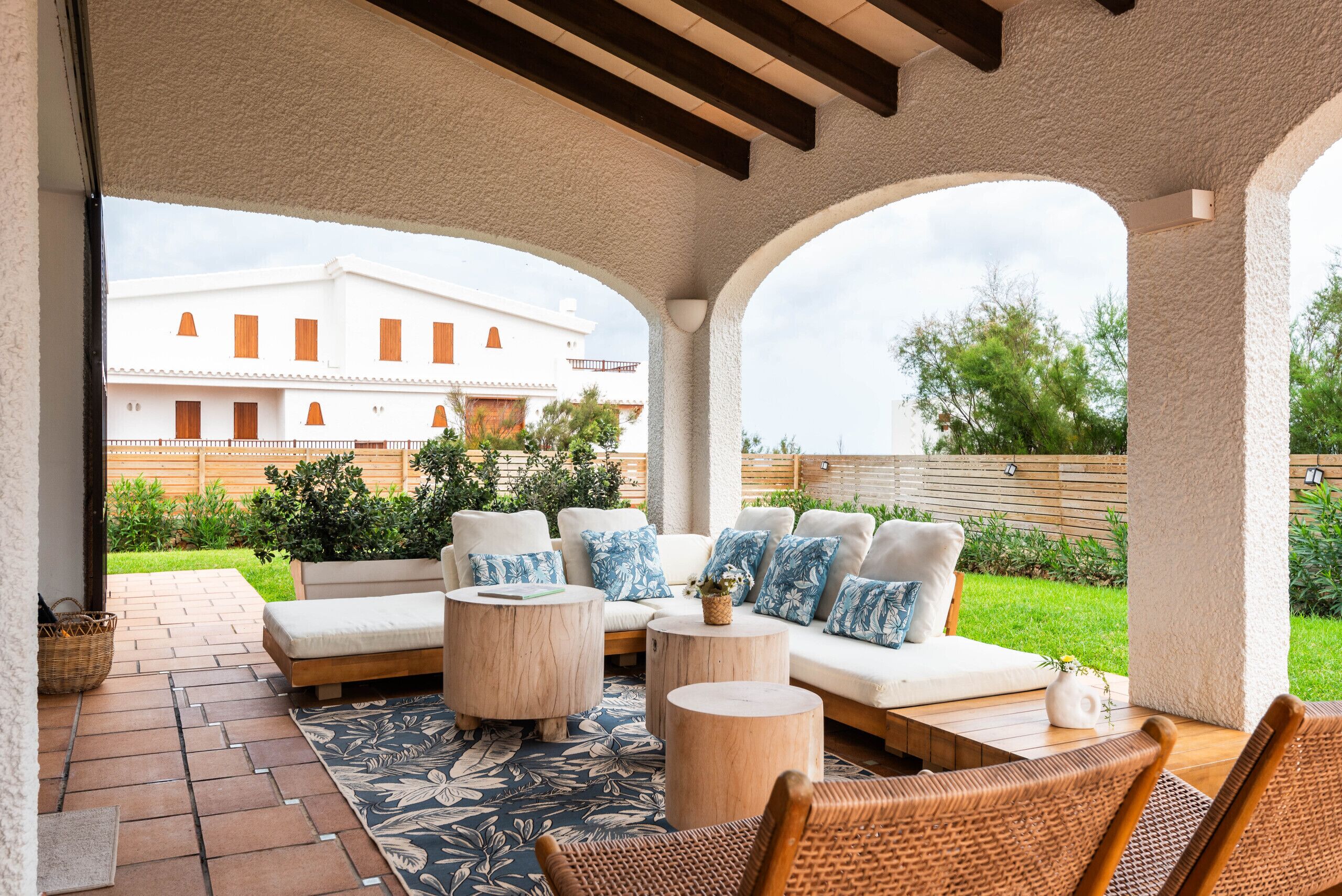 A covered outdoor seating area with a sofa and chairs, a patterned rug, and a view of a white building in the background.