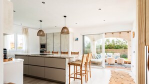 A modern kitchen with a large island, wooden chairs, and a view to an outdoor patio. - Kistine 5 bedroom villa, Cala Morell (Cala Morell)