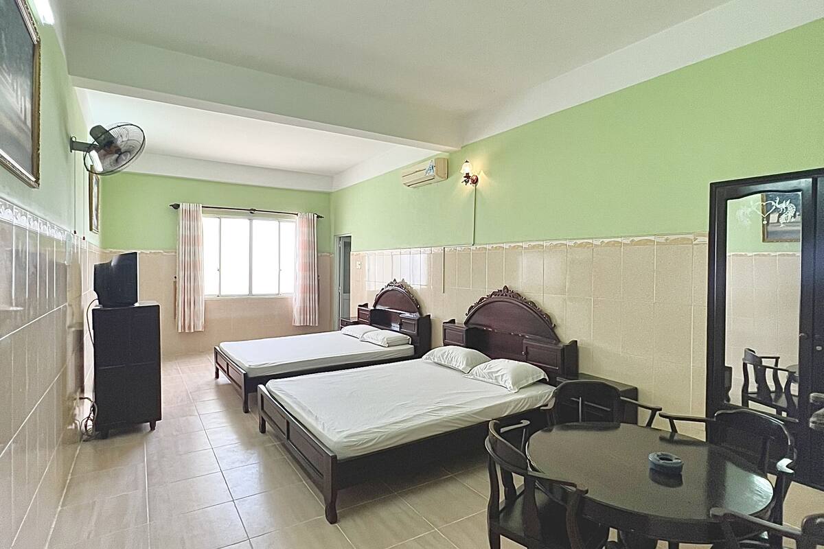 A hotel room with two beds, a dining table, a chair, a fan, a mirror, and a window.