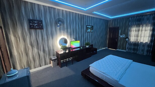 A bedroom with a bed, a desk with a computer, a chair, a clock, a mirror, and a wall with curtains. - Phi Long 2 Hotel Thu Dau Mot (Ho Chi Minh City)