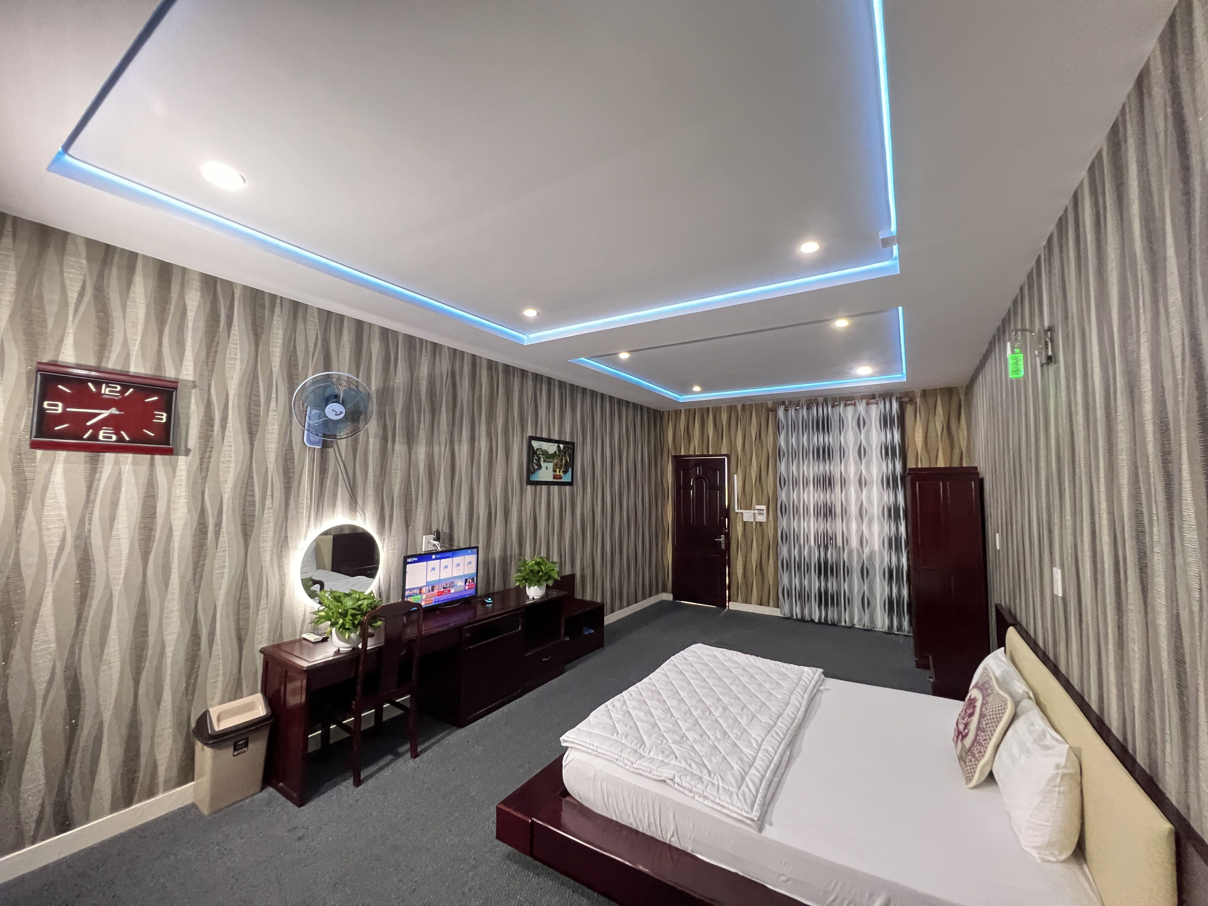 A modern bedroom with a bed, a desk, a computer, a clock, a fan, and a door.