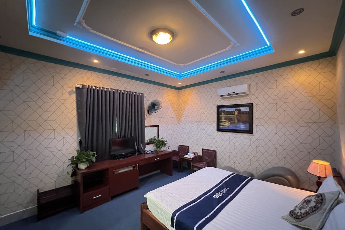 A hotel room with a bed, a desk, a chair, a TV, a fan, and a window with curtains.
