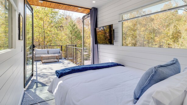 A bedroom with a bed, a TV, and a balcony with a sofa and railing. - New - Pine View MicroCabin - Rare (Broken Bow)