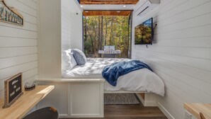 A bedroom with a built-in bed, a small table, a chair, and a window with a view of trees. - New - Pine View MicroCabin - Rare (Broken Bow)