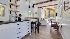 A modern kitchen with white cabinets, wooden beams, and a black countertop. There are two bar stools and a wooden table. - New - Pine View MicroCabin - Rare (Broken Bow)