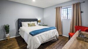 Une chambre à coucher avec un lit, une table de chevet, une lampe, une fenêtre avec des rideaux, une chaise rouge et une table en bois.