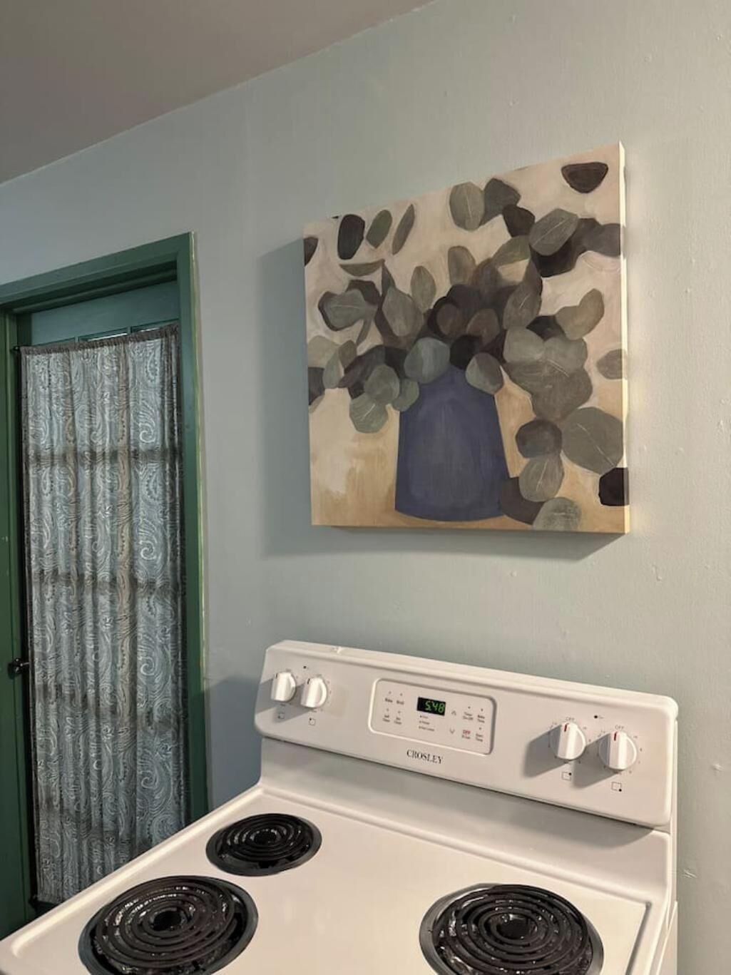 Een keuken met een witte Crosley-oven, een groene deur en een schilderij van een vaas met bladeren aan de muur.