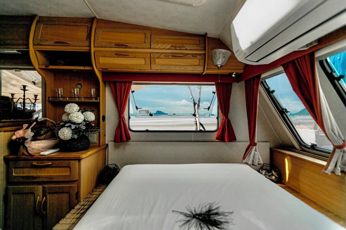 A cozy RV interior with a bed, wooden cabinets, a small table with flowers, and a window offering a beach view.