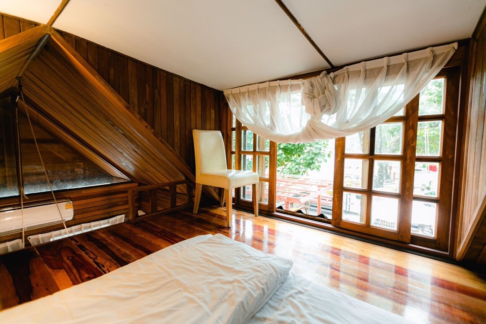 A room with a sloped ceiling, a bed, a chair, and large windows with curtains.