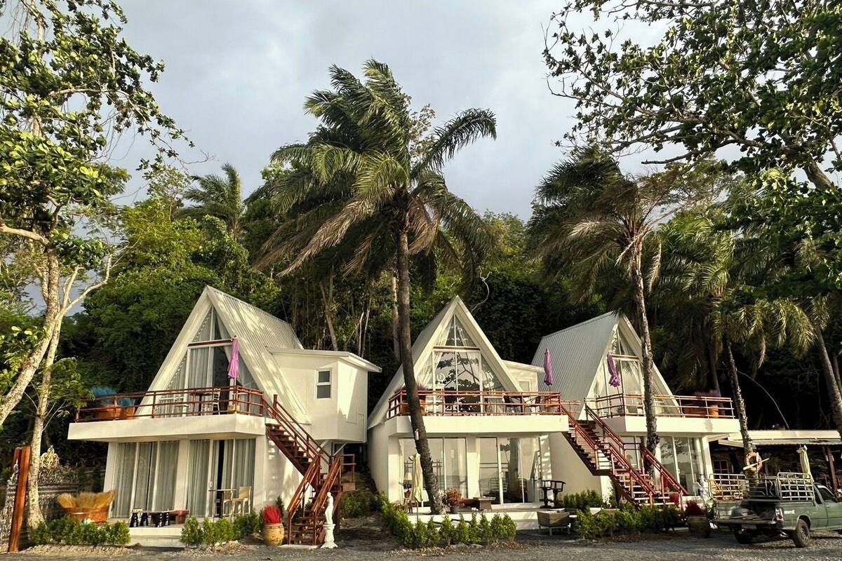 A row of modern, white, two-story houses with large windows and balconies, surrounded by lush greenery and palm trees.