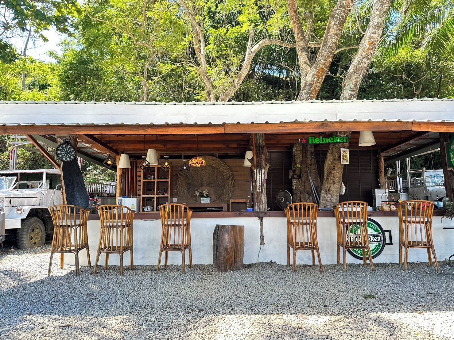 A rustic outdoor seating area with wicker chairs and a Heineken sign, surrounded by trees and a gravel ground.
