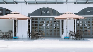 Exterior de un hotel con dos zonas de estar al aire libre, cada una con un paraguas con la palabra 'COFFEE' y sillas de metal.