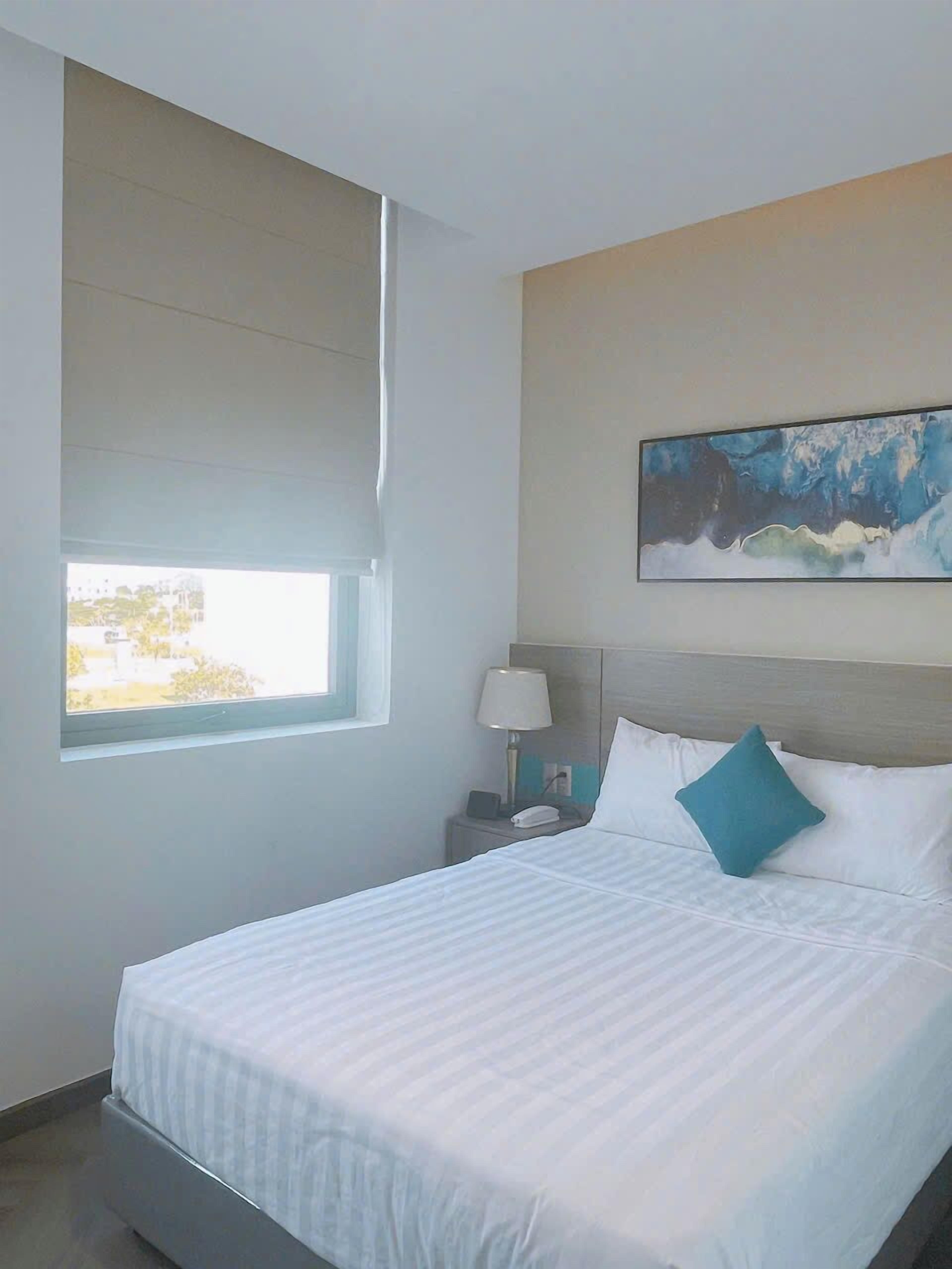 A hotel room with a large bed, a nightstand with a lamp, a painting on the wall, and a window providing a view.