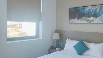 A hotel room with a large bed, a nightstand with a lamp, a painting on the wall, and a window providing a view.