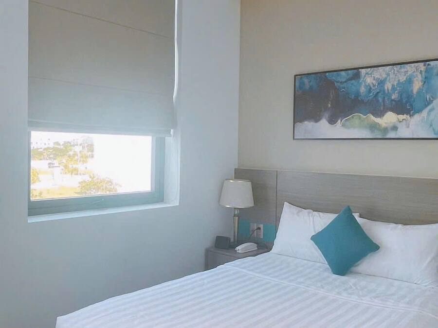 A hotel room with a large bed, a nightstand with a lamp, a painting on the wall, and a window providing a view.