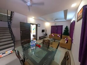 A modern living room with a glass dining table, a sofa set, a TV, and a staircase. - Atharva Villa (Lonavala)