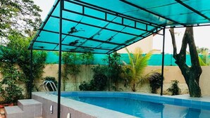 Ein Schwimmbecken mit Glasüberdachung, umgeben von einer gefliesten Terrasse und viel Grün.