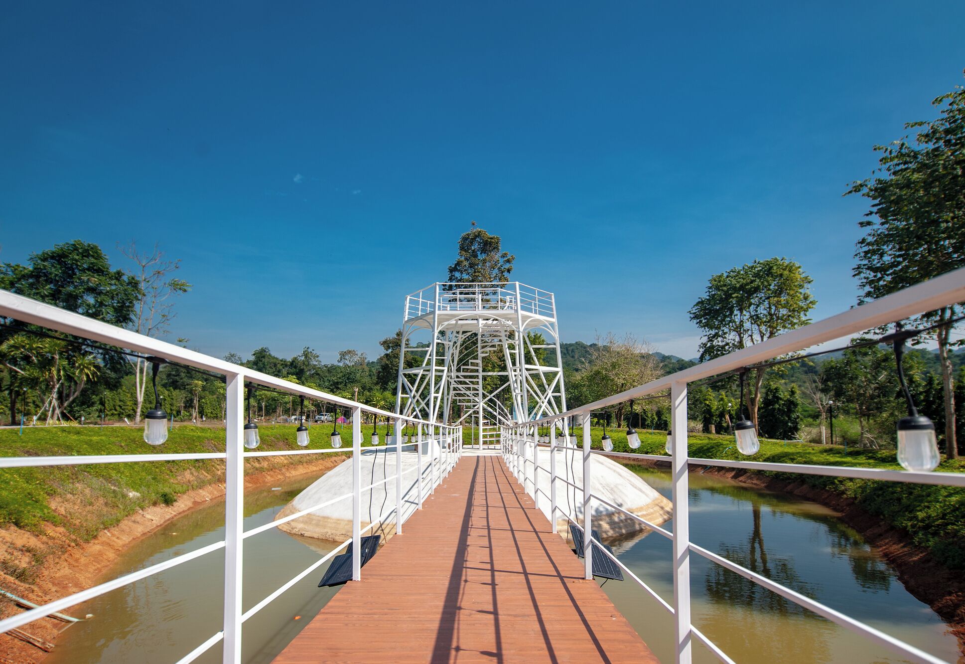 A wooden bridge with white railings leading to a white metal structure with stairs.