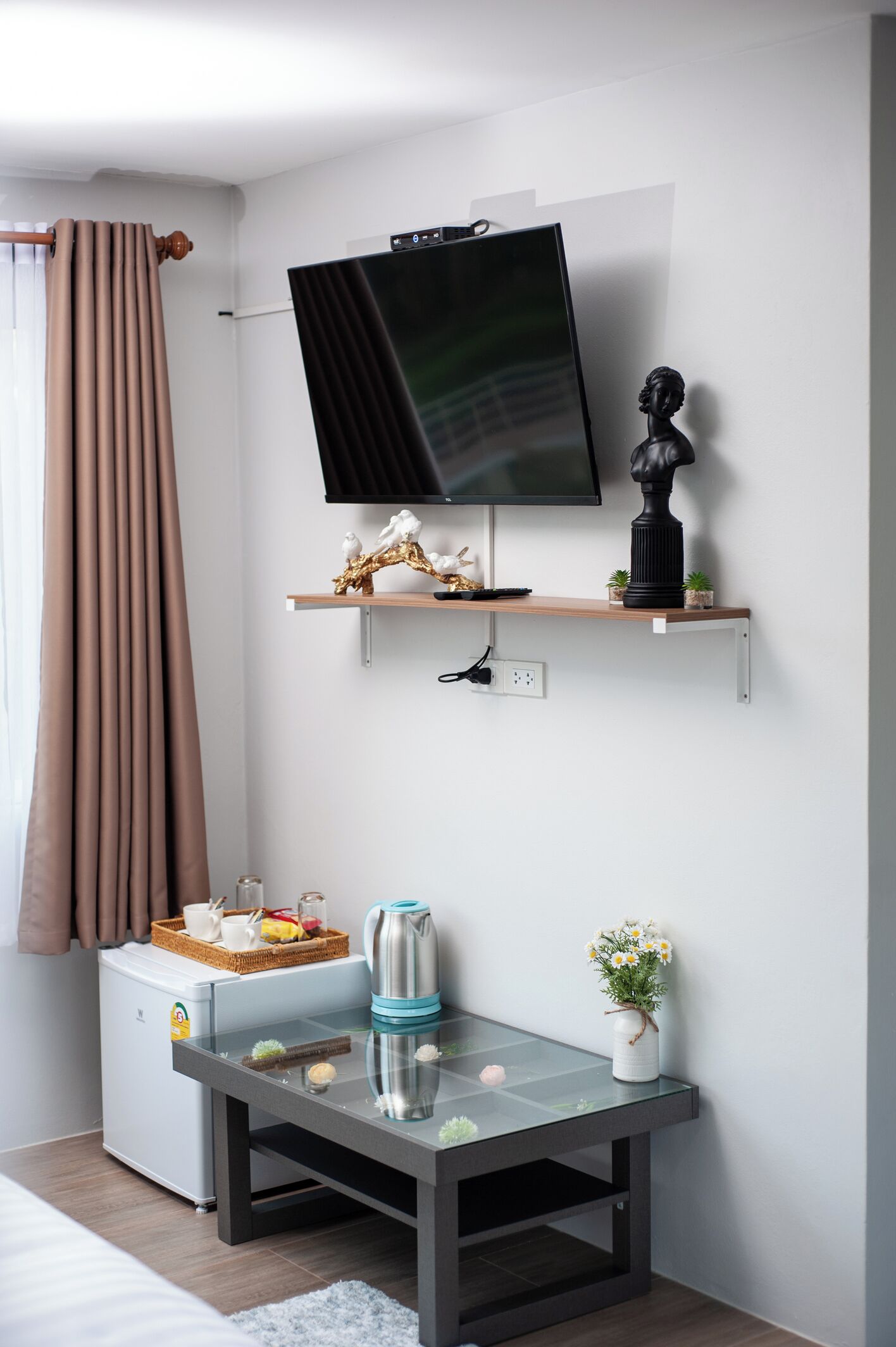 Una sala de estar moderna con un televisor de pantalla plana montado en la pared, una mesita con adornos, un refrigerador compacto y una ventana con cortinas.