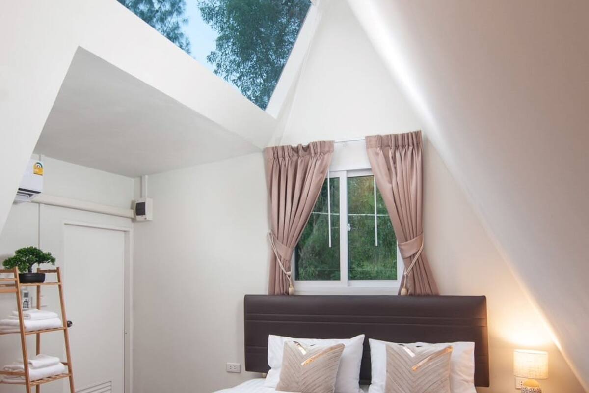 Une chambre avec un puits de lumière, un lit avec une literie blanche, une table de chevet avec une lampe et une fenêtre avec des rideaux.