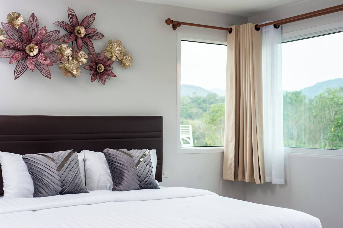 Une chambre à coucher avec un lit, des œuvres d’art décoratives au mur, une fenêtre avec des rideaux et une vue sur la verdure.