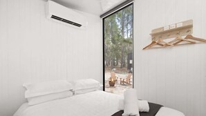 A bedroom with a bed, white walls, a window, and a view of an outdoor area with a fire and chairs. - Bilby Park Escape 1 by Tiny Away (Black Range)
