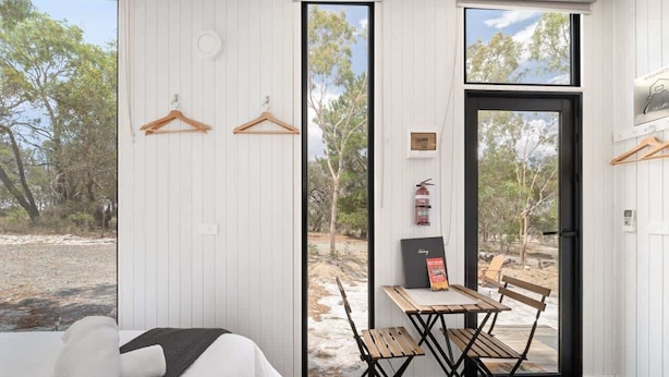 A room with a bed, a table with chairs, a fire extinguisher, and a view of the outdoors. - Bilby Park Escape 1 by Tiny Away (Black Range)