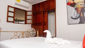 A bedroom with a bed, a wooden wardrobe, a mirror, a fan, and a painting of an elephant.