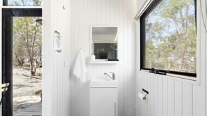 Un baño moderno con lavamanos, espejo y retrete blancos. Cuenta con una ventana que ofrece vistas a árboles y una terraza de madera en el exterior.