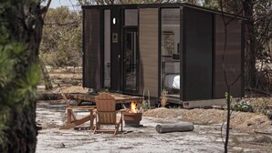 Una cabaña compacta y moderna con una pequeña terraza y una fogata, rodeada de vegetación natural.