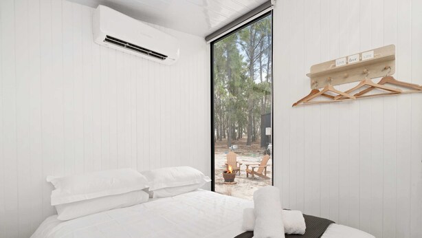 Un dormitorio con cama, ropa blanca, un tapete negro, una toalla, un panel de madera con letreros que dicen 'LIVE LOVE LIFE', y vistas a un espacio exterior con fuego y sillas.