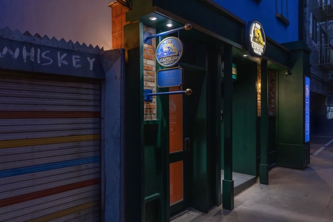 En byggnad med en stängd persienn på vänster sida och en skylt ovanför som visar ordet "WHISKEY". Ovanför ingången finns två upplysta skyltar, en med en tågliknande logotyp och den andra med en silhuett av en byggnad.