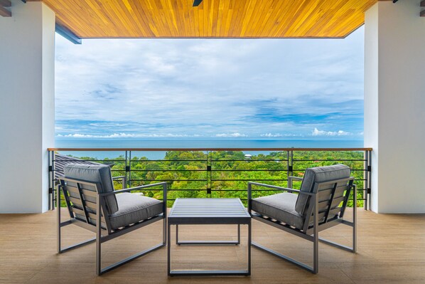A balcony with two chairs and a table, overlooking a lush green landscape and the sea.