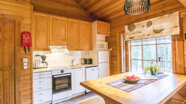 A wooden cabin kitchen with a large island, wooden cabinets, a microwave, a stove, and a window with a view of trees. - Terwa-ukko 2 by Interhome (Hyrynsalmi)