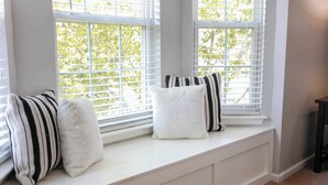 Un siège de fenêtre agrémenté de trois coussins, des murs blancs et une vue sur les arbres à l’extérieur.