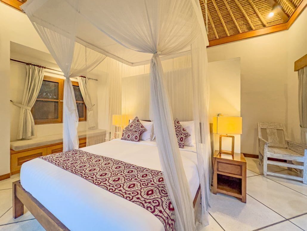 A four-poster canopy bed with a patterned bedspread, bedside lamps, a wooden nightstand, and a chair.