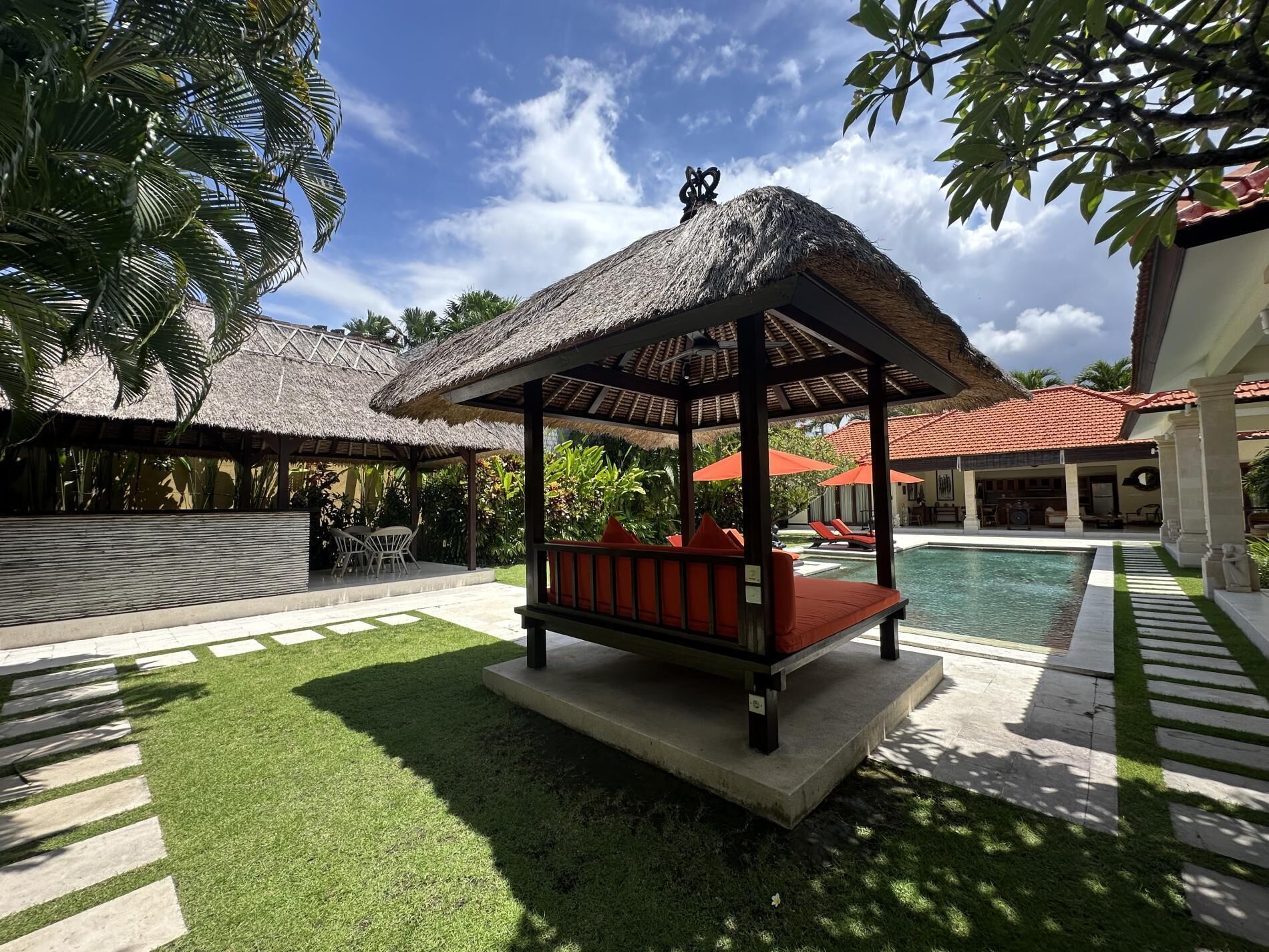 A thatched-roof gazebo with a bench, surrounded by a swimming pool and palm trees.