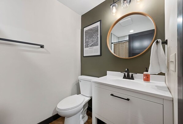 A modern bathroom with a white toilet, a white sink with a black faucet, a wooden-framed mirror, and a framed picture on the wall.