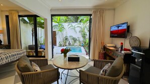 A hotel room with a bed, two armchairs, a small table, a TV, and a view of a pool and greenery. - Villa Bugis Kayu Aya Seminyak (Seminyak)