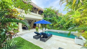 A poolside area with lounge chairs, a blue umbrella, and a thatched-roof building. - Villa Bugis Kayu Aya Seminyak (Seminyak)