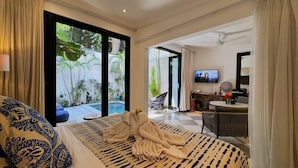 A bedroom with a bed, a TV, a desk, and a view of a pool and garden. - Villa Bugis Kayu Aya Seminyak (Seminyak)