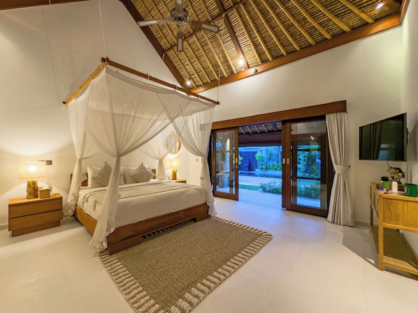 A bedroom with a canopy bed, a wooden nightstand, a bedside table with a lamp, a large window with curtains, and a view of the outdoors.