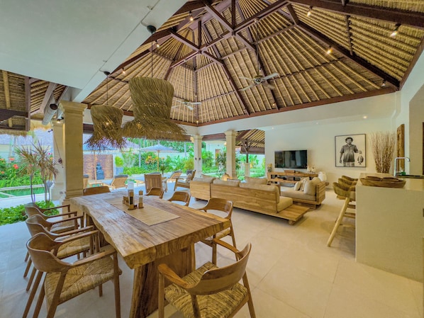A spacious living area with a wooden dining table, comfortable seating, a TV, and a large window overlooking a garden.