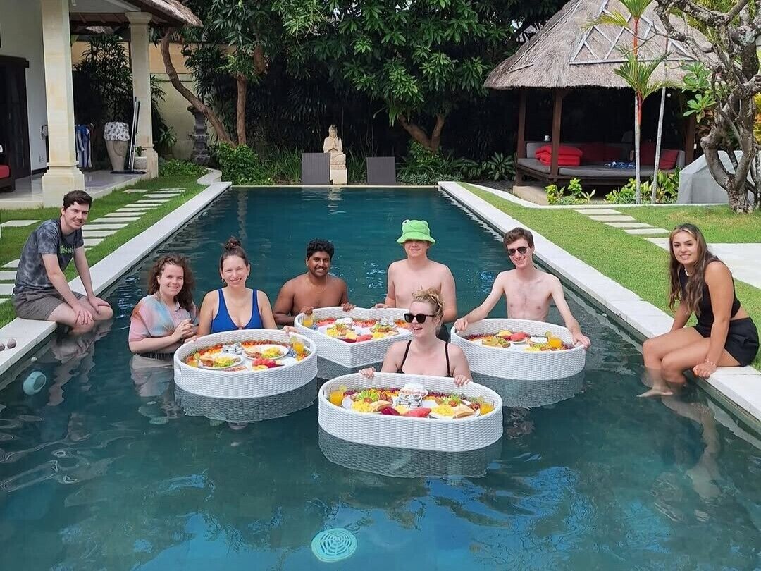 A pool area with people seated in circular wicker tables, surrounded by a well-maintained garden and a thatched-roof structure.