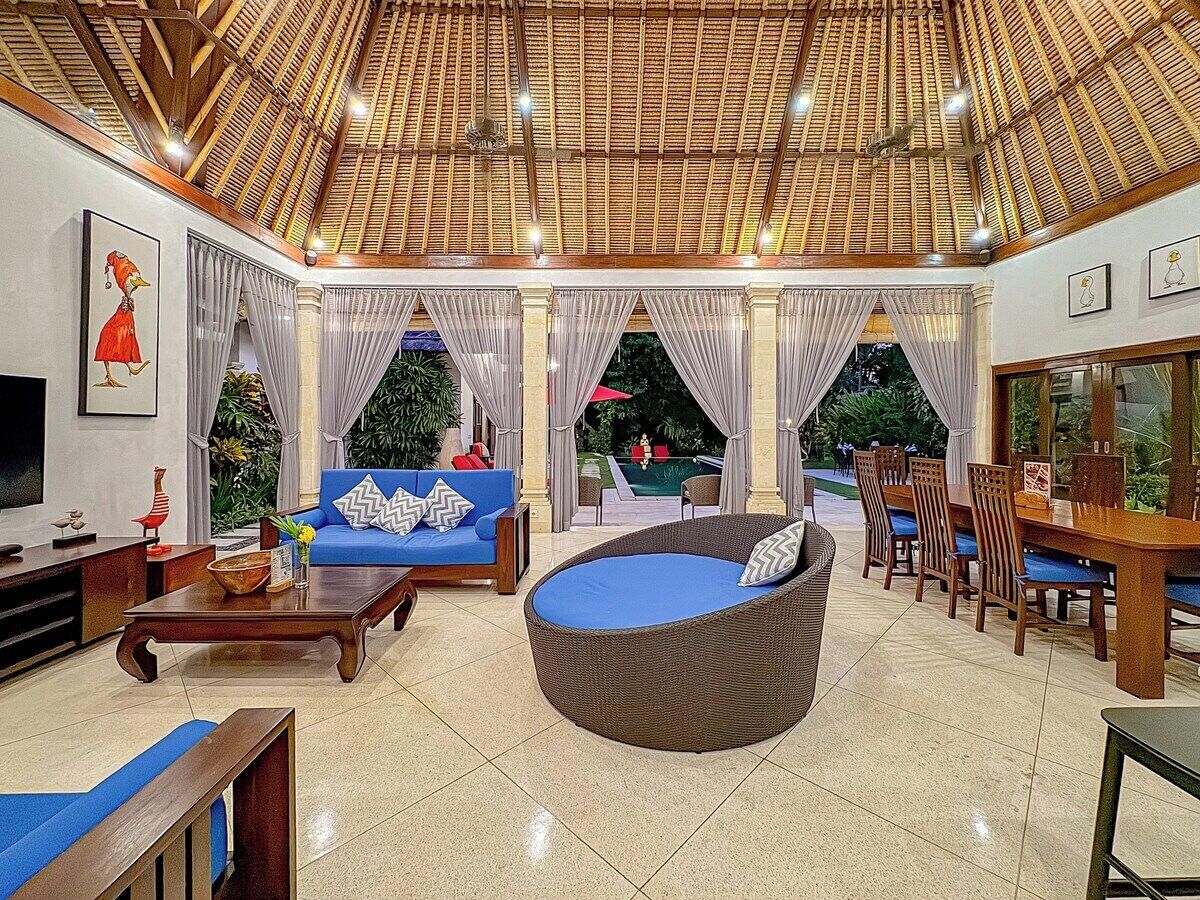 Un vaste espace intérieur doté d’un haut plafond, une table à manger avec des chaises, un canapé bleu, un fauteuil en osier et un téléviseur posé sur un meuble.