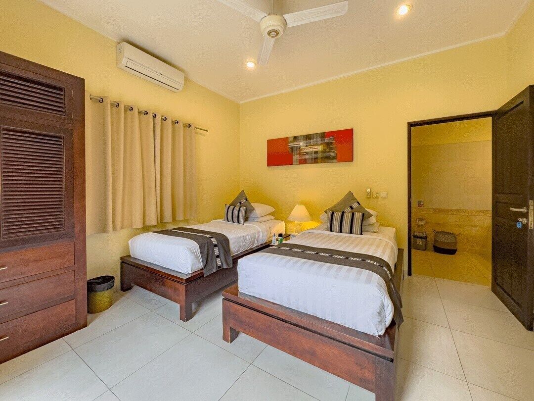 Une chambre d’hôtel avec deux lits, une armoire en bois, un climatiseur, un ventilateur de plafond, une salle de bain avec douche et un tableau au mur.